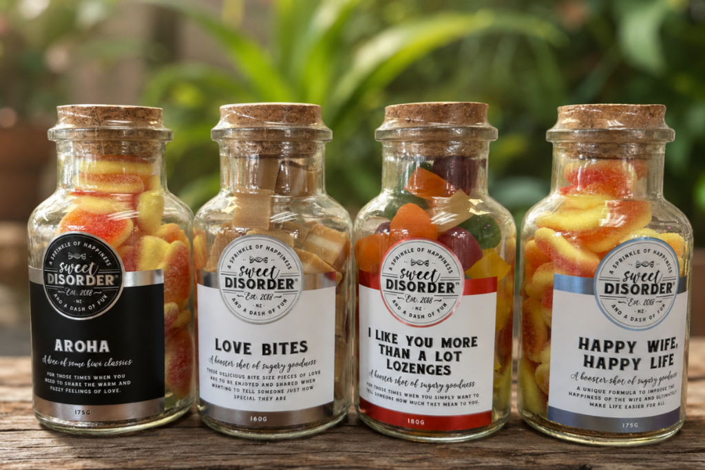 Four glass jars filled with colorful fruit and vegetable snacks, arranged on a rustic wooden surface with a blurred green garden background. Each jar has a cork lid and a white label with a different humorous slogan like 'AROHRA', 'LOVE BITES', 'I LIKE YOU MORE THAN A LOT LOZenges', and 'HAPPY WIFE HAPPY LIFE'.