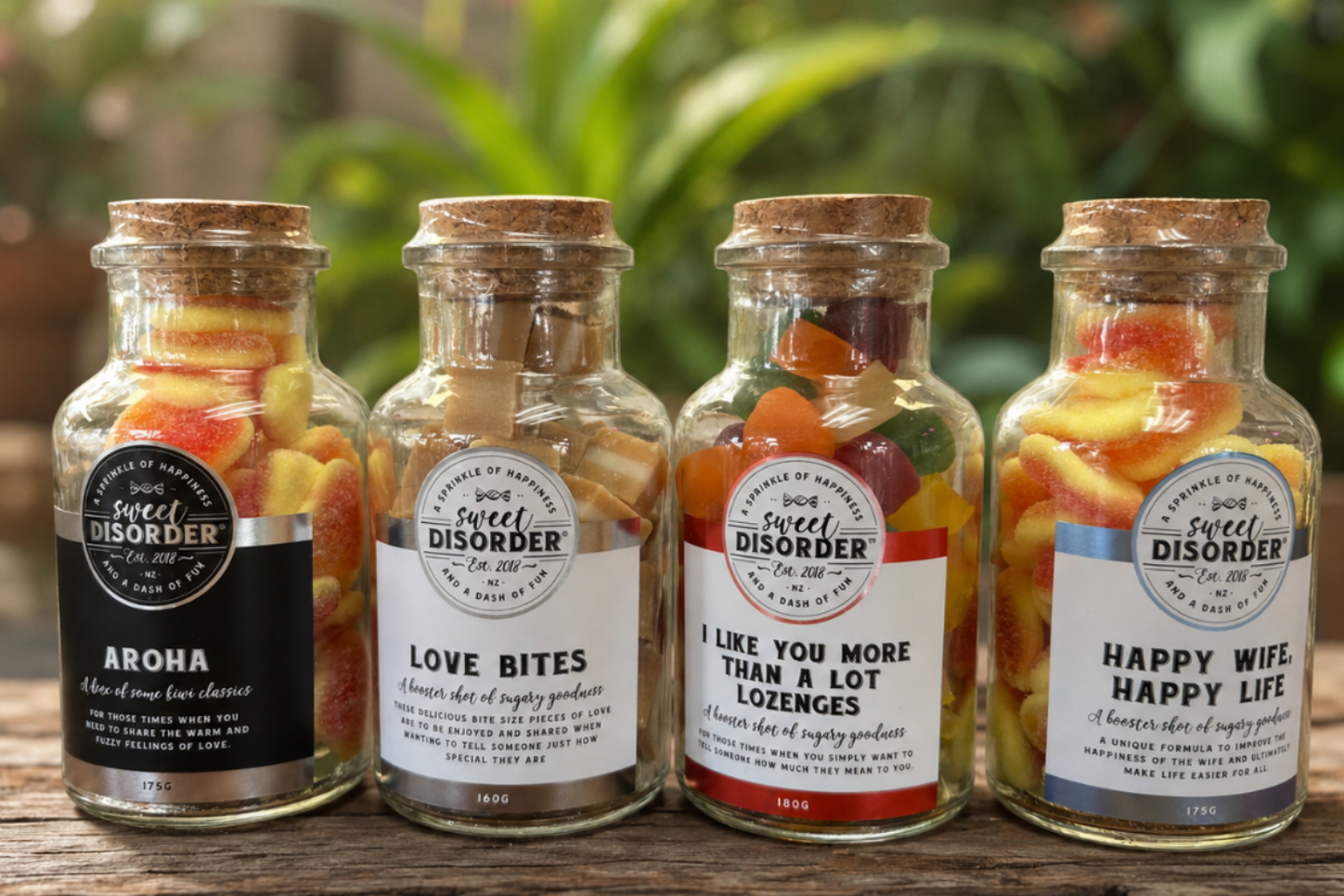 Four glass jars filled with colorful fruit and vegetable snacks, arranged on a rustic wooden surface with a blurred green garden background. Each jar has a cork lid and a white label with a different humorous slogan like 'AROHRA', 'LOVE BITES', 'I LIKE YOU MORE THAN A LOT LOZenges', and 'HAPPY WIFE HAPPY LIFE'.